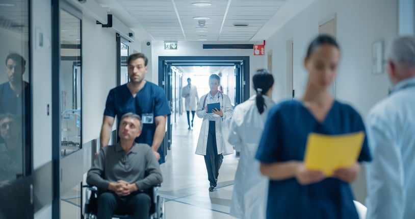 Valcom Life Safety AI Voice blog Busy Hospital Hallway Filled with Medical Experts in White Coats and Blue Uniforms. Middle Aged Female Doctor Reviewing a Patient's History on a Tablet Computer as She Heads to the Examination Room