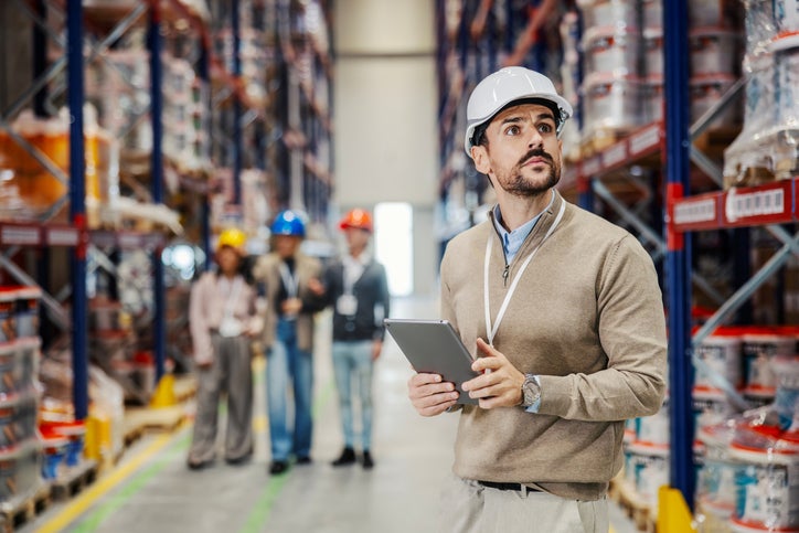 Male manager wearing a hard hat and holding a digital tablet, inspecting warehouse inventory with his team