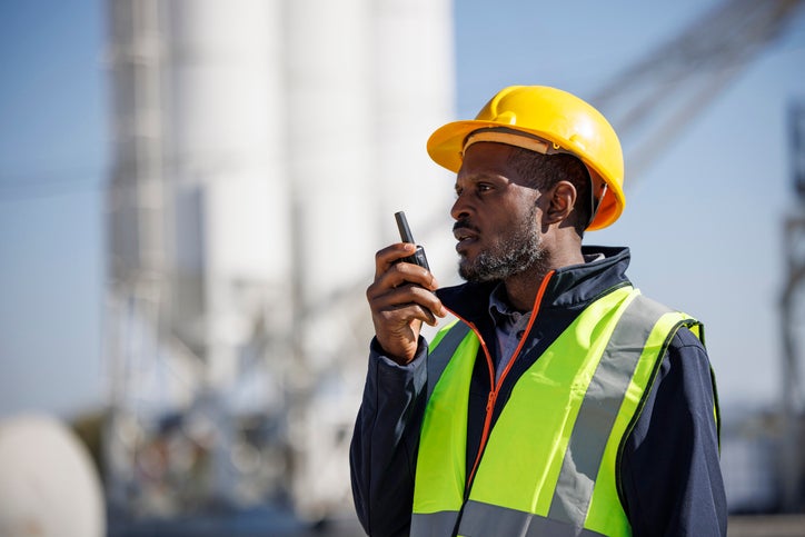 Valcom commerical industrial solutions image of an African American male in a yellow hard hat with a neon green safety vest speaking into a two-way radio outside an industrial plant